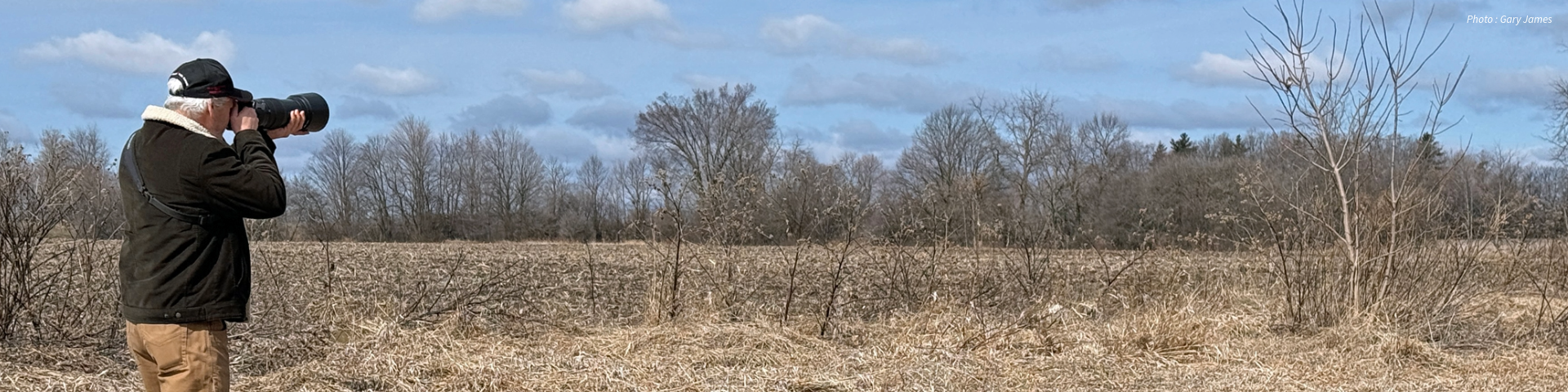 Une personne en veste noire et pantalon marron regarde à travers un grand objectif de caméra dans un champ, avec des arbres sans feuilles et un ciel bleu en arrière-plan.