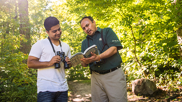 Un employé de Parcs Canada, vêtu d'un t-shirt vert, montre un livre à un randonneur en t-shirt blanc muni de jumelles. C'est une journée ensoleillée sur un sentier bordé d'arbres à la végétation luxuriante.
