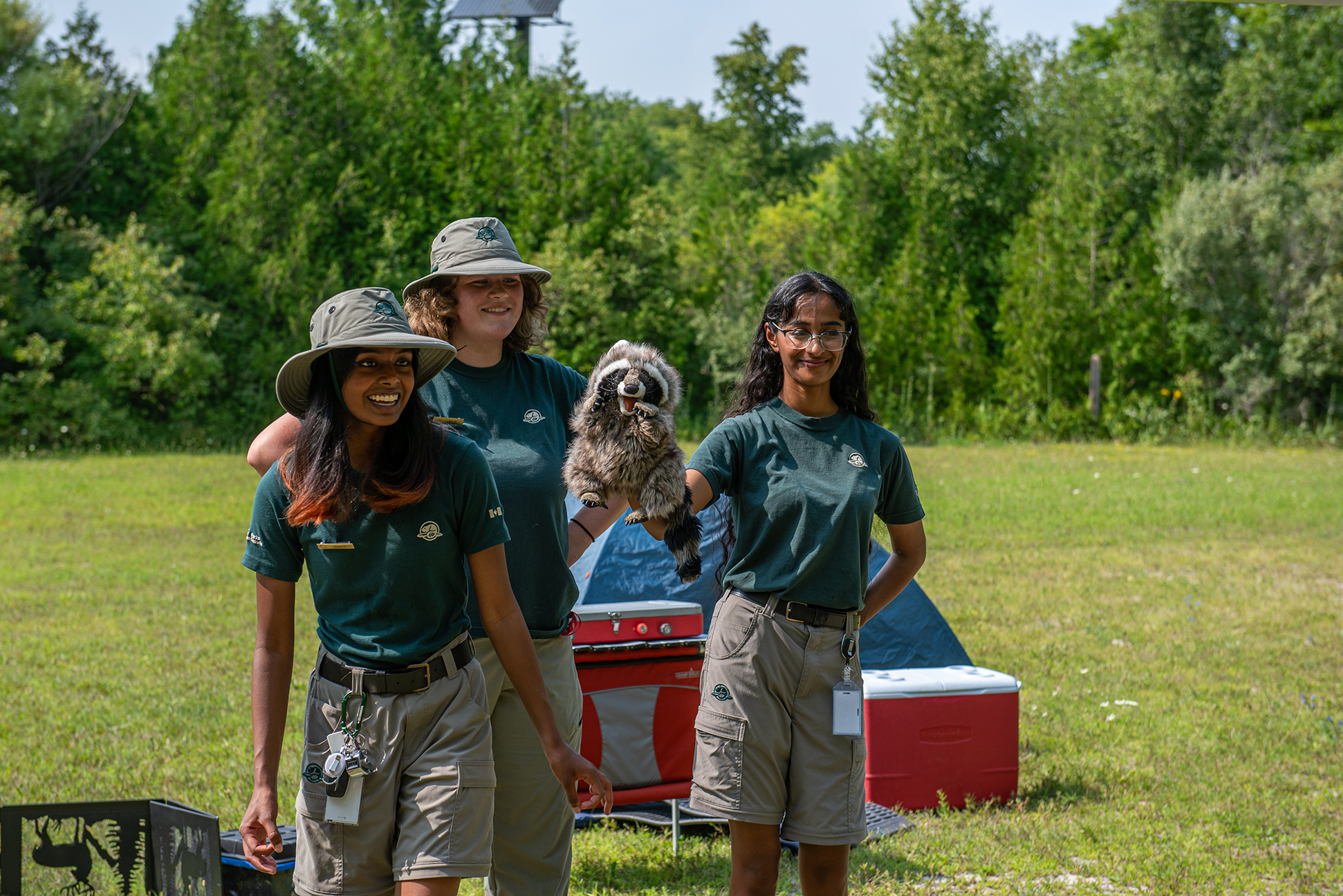 Three Parks Canada uniformed staff are smiling and standing in front of a camping site in the middle of a field on a sunny day. One staff member is holding a puppet in the shape of a raccoon. 
