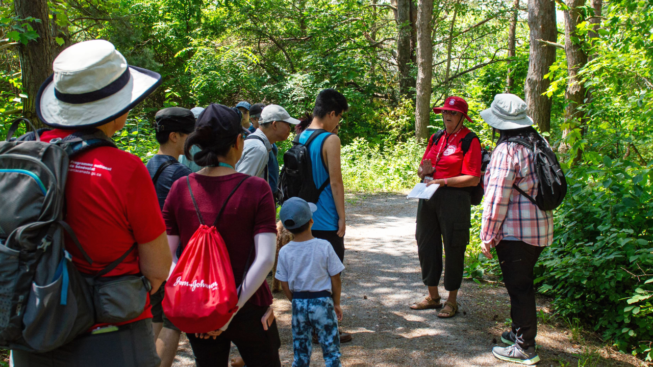 A group of people is participating in a guided walk at Rouge National Urban Park. The guide, wearing a red shirt and hat, stands on a gravel path surrounded by lush green trees and foliage. The participants are attentively listening, with some wearing backpacks and hats.