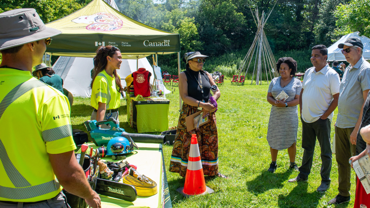A group of people interacts with park staff and Indigenous partners under a green tent labeled “Parks Canada” in Rouge National Urban Park. The tent displays safety gear and informational materials. Indigenous structures (tipi) and dense greenery are visible in the background.