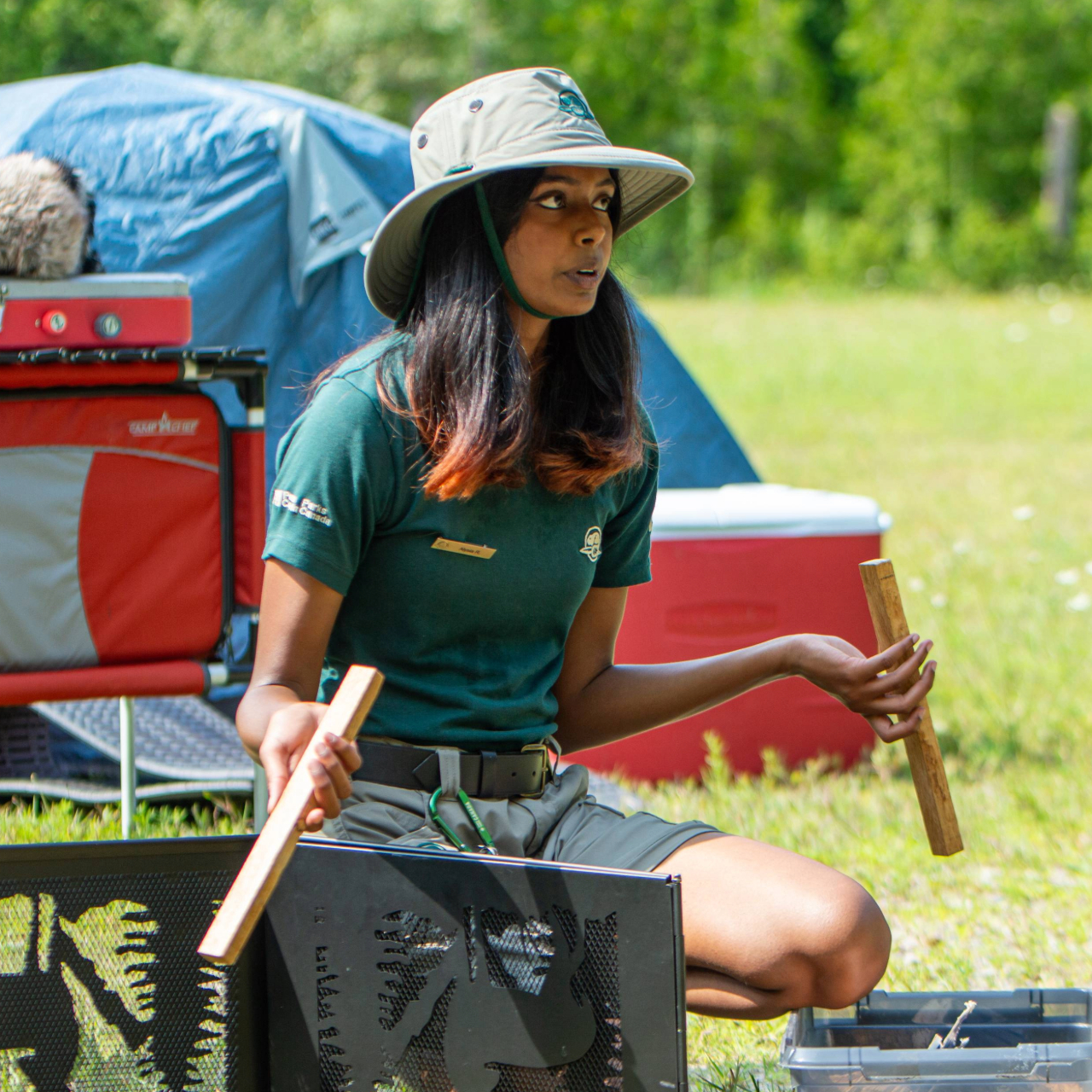 A Parks Canada staff member is giving a camping presentation outdoors. They are wearing a green Parks Canada shirt, beige shorts, and a wide-brimmed hat. The staff member is holding two wooden sticks and sitting on the grass next to a black metal fire pit with animal silhouettes. In the background, there is camping equipment including a blue tent, red cooler, and other supplies.