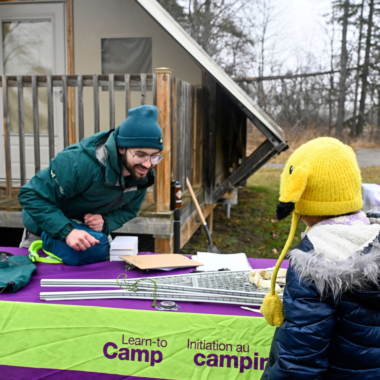 A Parks Canada staff member stands behind a table covered with a purple and green cloth featuring the Parks Canada logo and icons for camping, biking, hiking, and campfires. They are interacting with a child. The child is wearing a yellow hat with ear flaps and a blue winter coat. They are outdoors near a cabin with trees in the background.