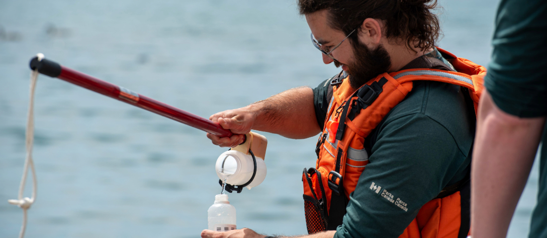 A person in an orange life jacket and green Parks Canada long-sleeve shirt collects a water sample at Rouge Beach using a sampling pole and white bottle. Lake Ontario is visible in the background.