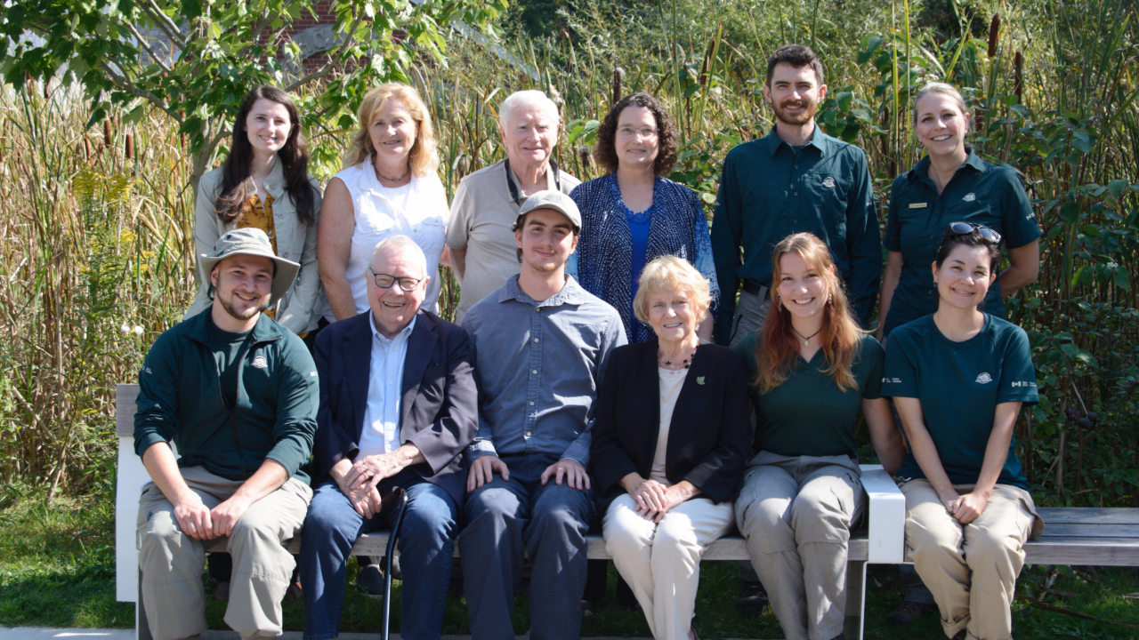 A group of 12 people posing outdoors in front of tall grasses and trees. Several individuals are wearing green Parks Canada uniforms, while others are dressed in casual or business attire. They are seated and standing on a bench in a natural setting.