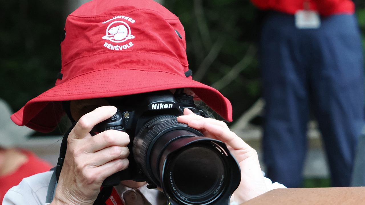 A person wearing a red hat with the text "VOLUNTEER BÉNÉVOLE" and an emblem is holding a Nikon camera and appears to be taking a photograph. Other people are visible in the background, including one wearing a red shirt and blue pants.