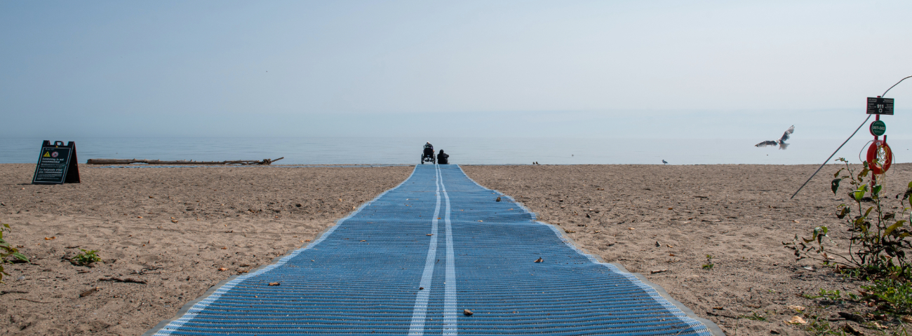 At Rouge National Urban Park, a blue Mobi Mat extends across a sandy beach toward Lake Ontario, enhancing accessibility. Two people sit at the end of the mat facing the water. A seagull flies nearby. A black info sign stands to the left; a lifebuoy and signage are on the right.