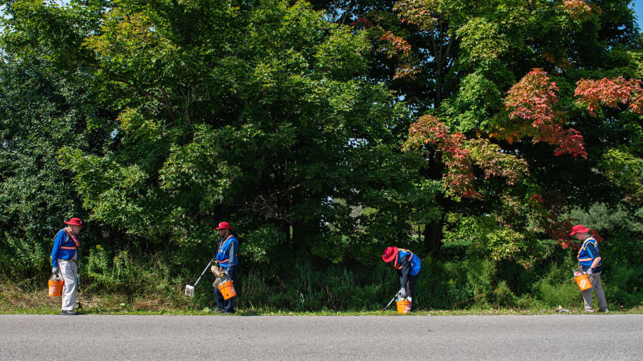 A group of four people wearing blue shirts and red hats are cleaning up the roadside at Rouge National Urban Park. They are holding orange buckets and using tools to pick up litter. The background consists of dense green trees with some leaves turning red, indicating the onset of autumn.