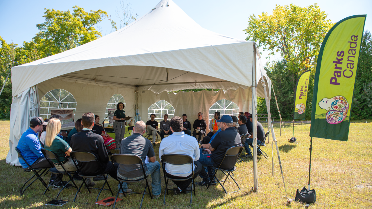 Group seated in a circle under a white tent at Rouge National Urban Park, with two Parks Canada flags in view.
