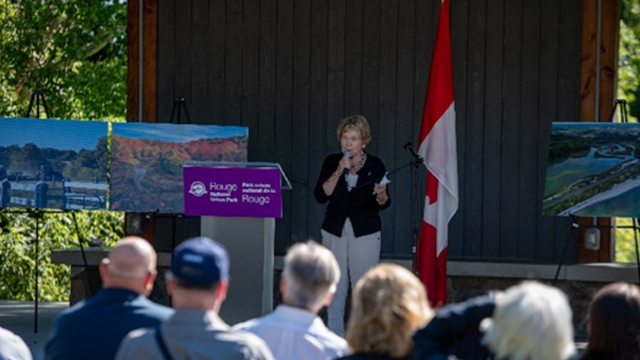 Speaker addressing an outdoor audience near a Canadian flag with landscape photos displayed on easels behind her.