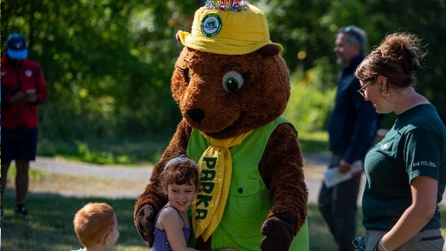 Person in a brown Parka costume wearing a yellow hat and green vest interacting with a small child outdoors on a sunny day.
