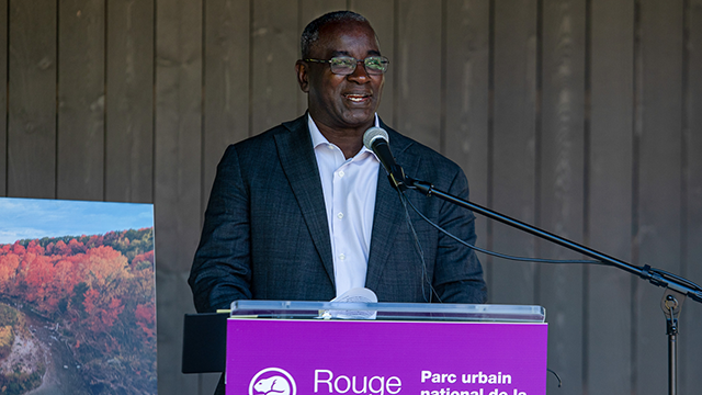 Man in a dark suit speaking at a podium with a purple sign reading 'Rouge Parc urbain naturel' and a microphone setup.