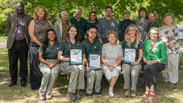 Group photo of fifteen people outdoors, with five seated in front holding certificates and others standing behind under green trees.