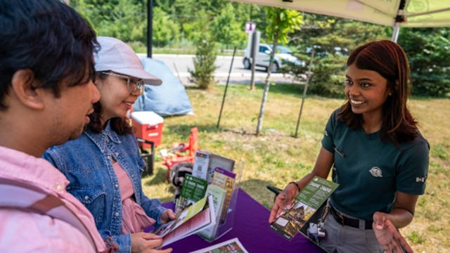 A Parks Canada staff member in a green shirt shows a brochure to two adult visitors under a yellow tent on a sunny day.