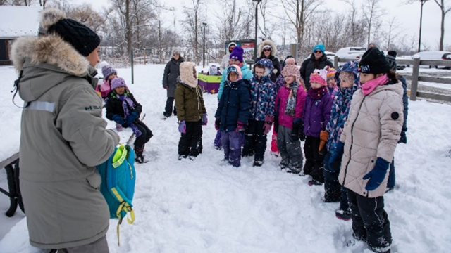 A Parks Canada staff member wearing a heavy beige parka talks to a large group of individuals standing in snow. 