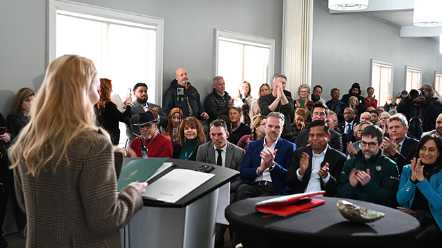 Speaker addressing a diverse audience seated and standing in a bright conference room with large windows.