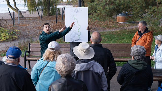 Un groupe en plein air écoute un employé de Parcs Canada qui explique un itinéraire tracé sur une grande carte posée à côté d'un banc, près de l'eau.