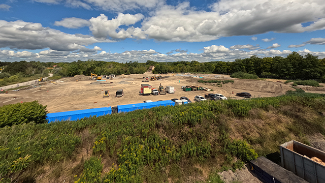 Wide view of a construction site with heavy machinery and vehicles under a partly cloudy sky, surrounded by greenery.