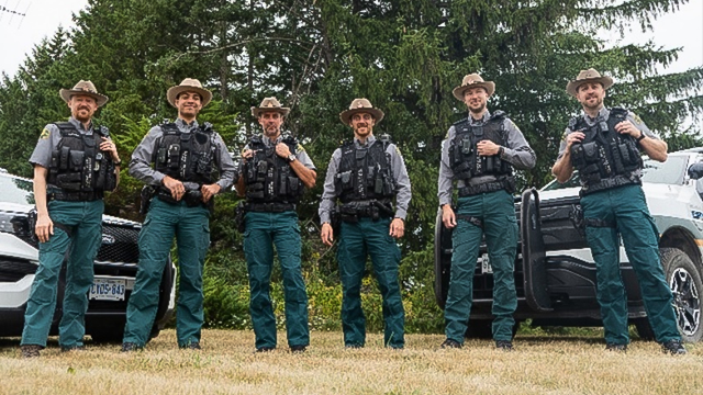 Six Parks Canada Wardens stand side by side posed for a photo in front of two parked white trucks and a large green tree.