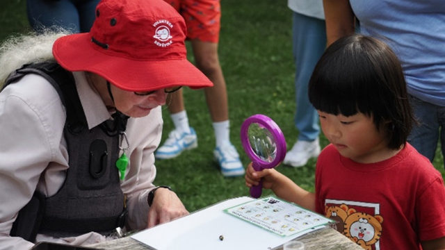 Young girl in red shirt examining a small object on a white table with a purple magnifying glass outdoors. Guided by an adult wearing a red sunhat and white shirt.
