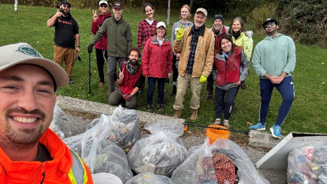 Group of volunteers posing outdoors behind collected trash bags during a cleanup event on a grassy area.