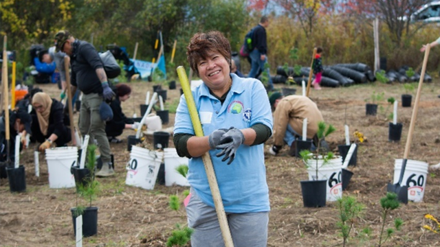 Group of volunteers planting young trees in a community garden on a clear day, with one person in a blue shirt holding a wooden stake in the foreground.