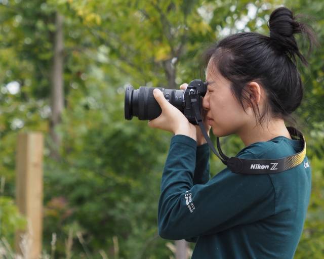 A person with their hair in a bun is taking a photo with a Nikon Z camera in a green outdoor setting.