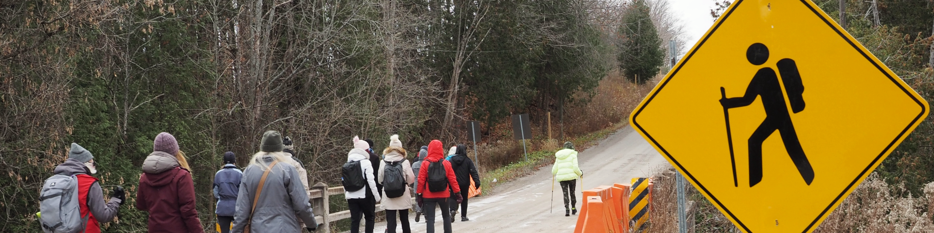 Un groupe de personnes vêtues de vêtements d'hiver marchent le long d'une route dans le parc urbain national de la Rouge. La route est entourée d'arbres aux branches nues. Sur le côté droit de l'image, il y a un panneau routier jaune avec une silhouette noire d'une personne faisant de la randonnée avec un sac à dos et un bâton de marche.