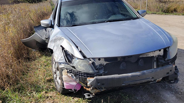 An old silver car that has been smashed up sits by a road surrounded by farm fields