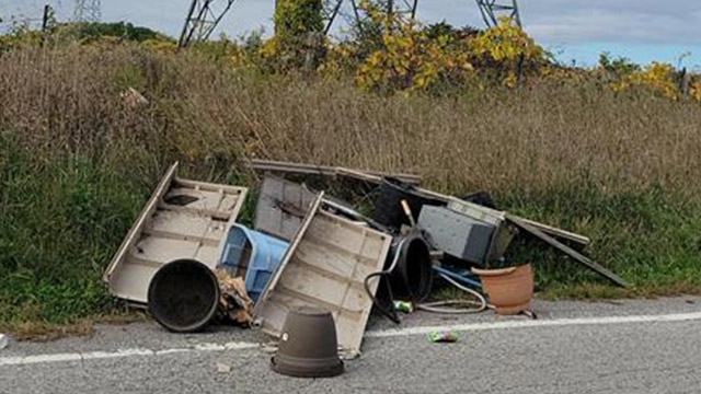 Large pieces of trash, old flower pots, wood, and other garbage items lie in a pile beside a road surrounded by long grass. 