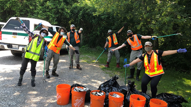 Six Parks Canada staff with florescent orange and yellow vests, stand along side a road with collected garbage bags and orange buckets filled with trash. 