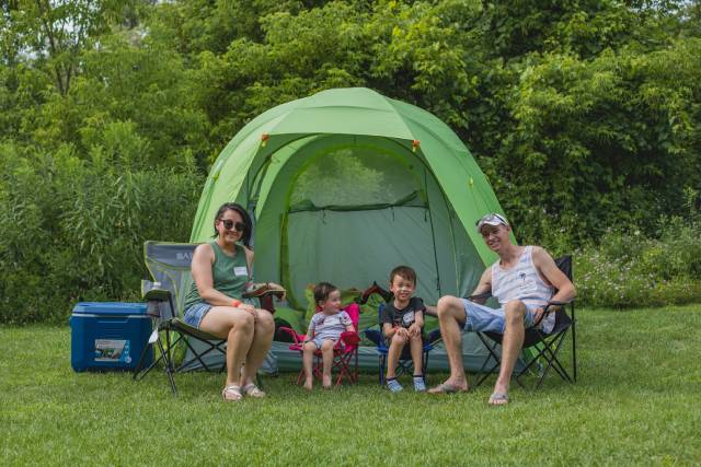 A family of four is sitting in front of a green tent outdoors