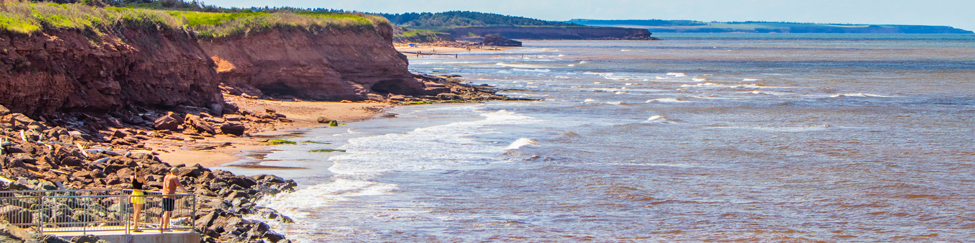 Two visitors stand on a look out viewing high red cliffs, and wavy waters.