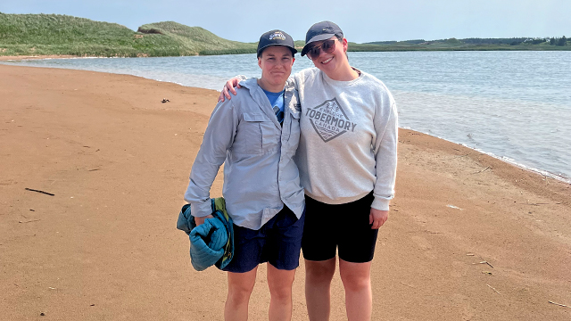 Dr Libby George and former Masters student Brianna Lunardi, stand side by side on the beach with lush green dunes in the background and a clear blue sky. 