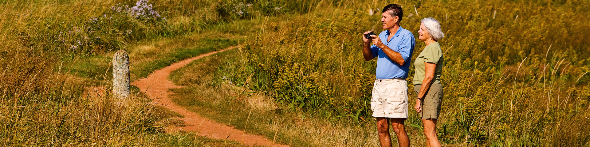 Two seniors stand along a trail in the park holding binoculars.