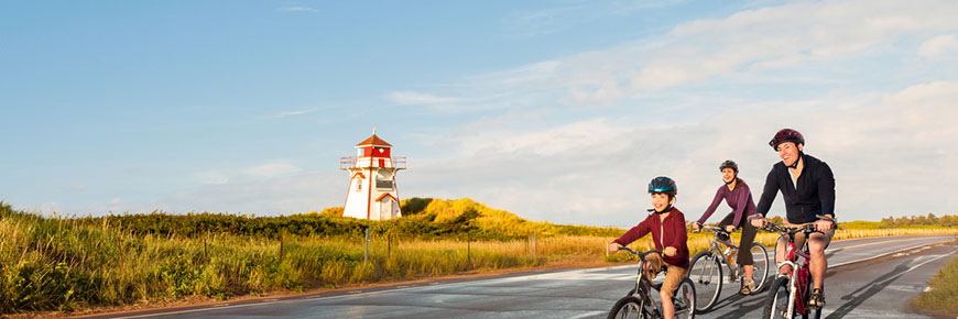 Two adults and a child, all wearing helmets, cycle past the Covehead Lighthouse on a sunny day. 