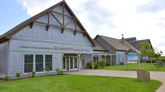 Greenwich Interpretation Centre is a large building with cedar siding and brown roof.  The grass is green and there is blue skies with fluffy white clouds.
