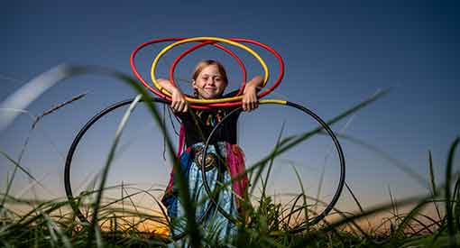 A young girl holds several hoops in many colours in tall grass as the sun sets