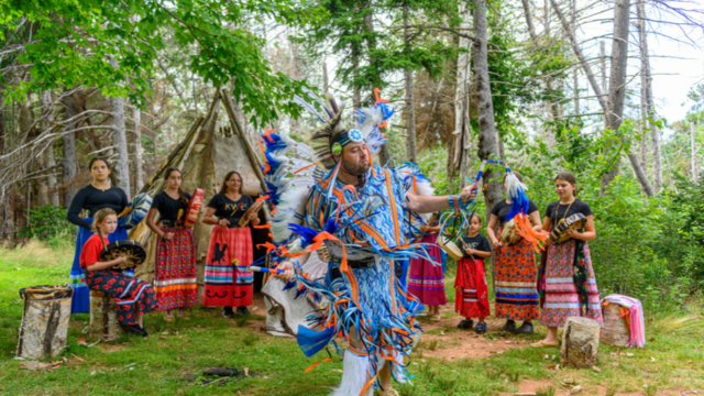 Un adulte vêtu d'un costume aux couleurs vives, bleu et orange, exécute une danse traditionnelle en plein air, entouré d'autres adultes et de quelques jeunes en tenues colorées, tenant des tambours.