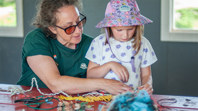 A Parks Canada staff member wearing a green shirt, helps a young girl wearing a beach hat at a table covered in yarn.