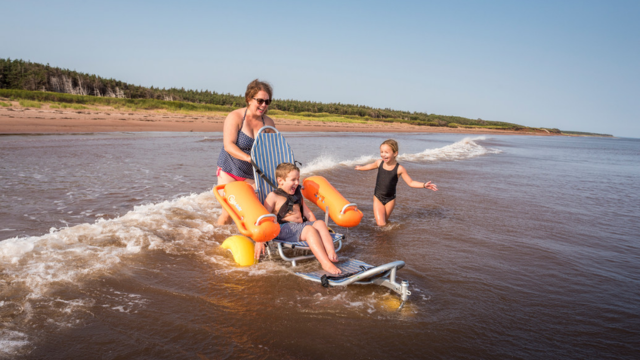 Une famille présentant des besoins en matière d’accessibilité nage dans l’océan en prenant place sur un fauteuil roulant à la plage Stanhope. Parc national de l’Île-du-Prince-Édouard.