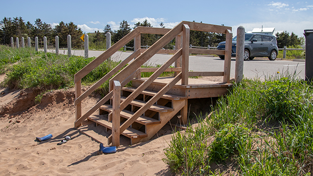 Escalier en bois menant d'une plage de sable à un parking pavé, avec une voiture garée à proximité.