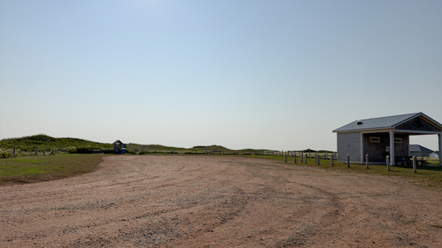 Une vaste étendue de gravier, avec un petit bâtiment gris sur la droite et des dunes herbeuses au loin, sous un ciel bleu limpide.