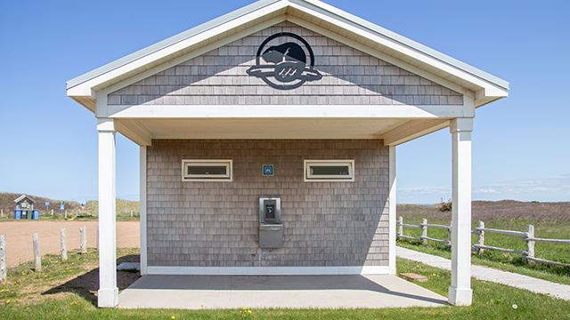 Petit pavillon en bord de mer, avec une cabine téléphonique fixée à un mur recouvert de bardeaux, sous un toit à deux versants, par une journée ensoleillée.
