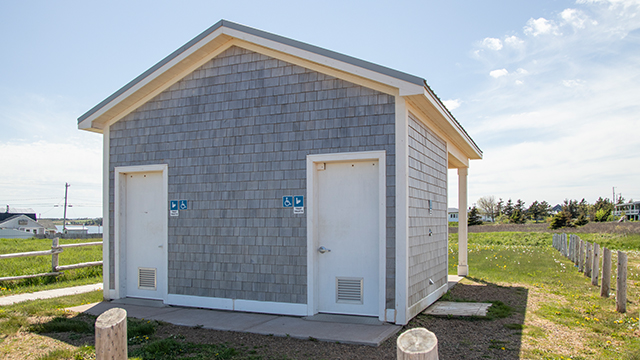 Petit bâtiment gris abritant des toilettes publiques, doté de deux portes blanches et de panneaux « accès handicapés » sur chacune d'elles, situé dans un espace ouvert et verdoyant sous un ciel dégagé.