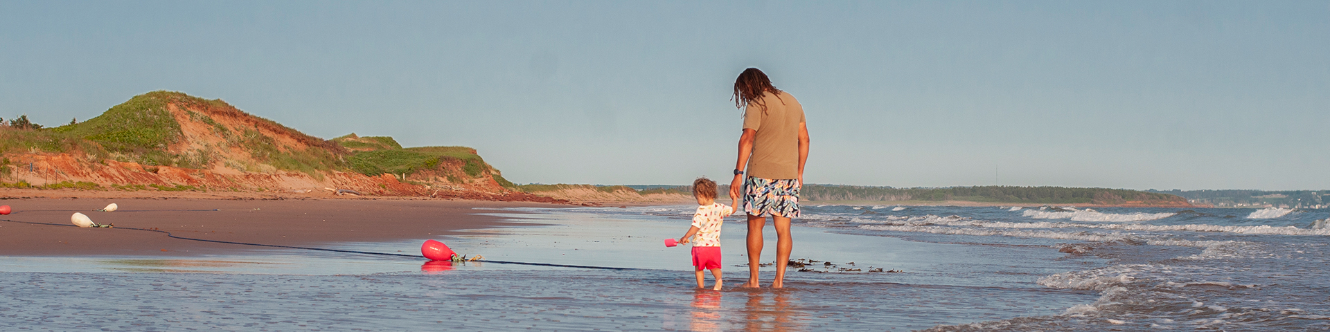 An adult walks along the waters edge with a small child.  There is grass covered sand dunes and a bright blue sky. 