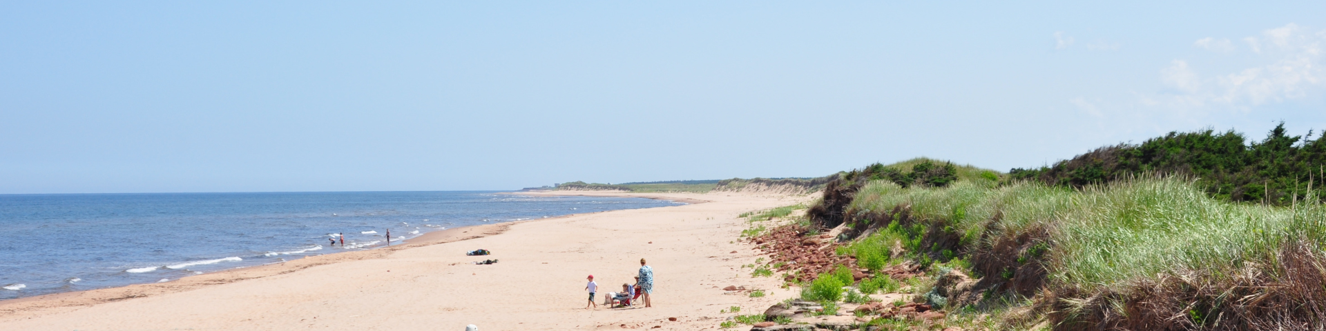 Several visitors are off in the distance at a white sandy beach with grass covered dunes, dark blue water and a bright blue sky.