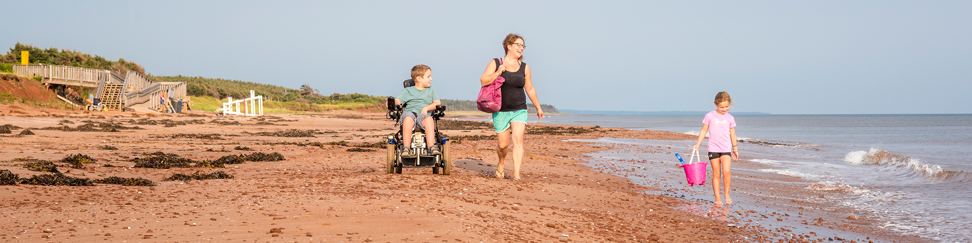 A mother walks along Stanhope Maine beach.  A young girl walks in the water with a sand bucket.  A young boy moves along in a beach wheelchair. 