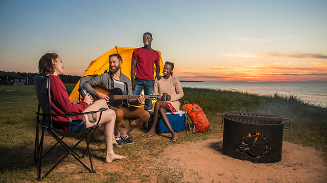 Four young adults sit around a campfire near the beach at sunset.