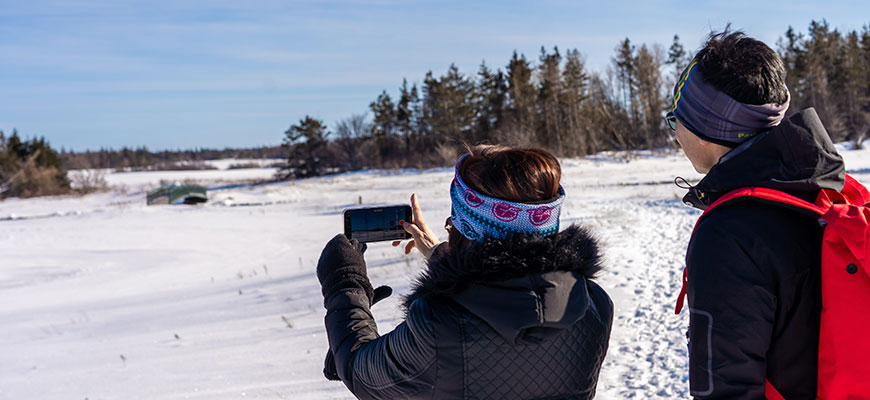 Two hikers take a photo with a cell phone on a bright winter day in PEI National Park on a snowy trail. 